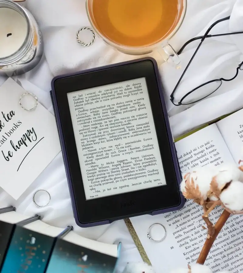 A cozy flat lay of an ebook reader with a cup of tea, open books, cotton flowers, rings, and glasses on a white background — perfect for book lovers and digital reading.